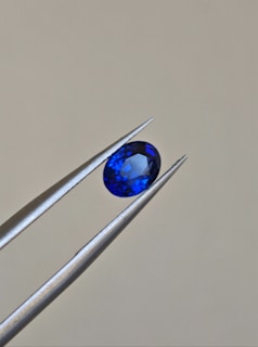 A pair of metallic tweezers delicately holds a small, oval-cut blue gemstone against a neutral background. The gemstone displays a vibrant blue color with facets that catch the light.