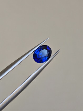 A pair of metallic tweezers delicately holds a small, oval-cut blue gemstone against a neutral background. The gemstone displays a vibrant blue color with facets that catch the light.