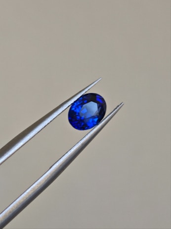 A pair of metallic tweezers delicately holds a small, oval-cut blue gemstone against a neutral background. The gemstone displays a vibrant blue color with facets that catch the light.