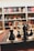 A close-up of a chessboard with black and white chess pieces arranged for play. The background consists of bookshelves filled with various books, creating a cozy, intellectual atmosphere.