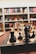 A close-up of a chessboard with black and white chess pieces arranged for play. The background consists of bookshelves filled with various books, creating a cozy, intellectual atmosphere.
