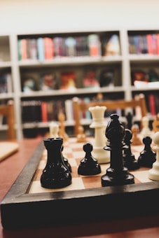 A close-up of a chessboard with black and white chess pieces arranged for play. The background consists of bookshelves filled with various books, creating a cozy, intellectual atmosphere.