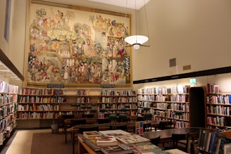 A well-lit library room with numerous bookshelves filled with books. In the center of the image, a large, colorful mural depicting various historical or cultural scenes covers the upper part of the wall. Several wooden tables and chairs are arranged neatly in the middle of the room. A modern light fixture hangs from the ceiling, casting warm light over the area.