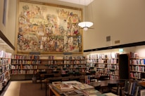 A well-lit library room with numerous bookshelves filled with books. In the center of the image, a large, colorful mural depicting various historical or cultural scenes covers the upper part of the wall. Several wooden tables and chairs are arranged neatly in the middle of the room. A modern light fixture hangs from the ceiling, casting warm light over the area.