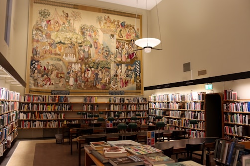 A well-lit library room with numerous bookshelves filled with books. In the center of the image, a large, colorful mural depicting various historical or cultural scenes covers the upper part of the wall. Several wooden tables and chairs are arranged neatly in the middle of the room. A modern light fixture hangs from the ceiling, casting warm light over the area.