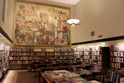 A well-lit library room with numerous bookshelves filled with books. In the center of the image, a large, colorful mural depicting various historical or cultural scenes covers the upper part of the wall. Several wooden tables and chairs are arranged neatly in the middle of the room. A modern light fixture hangs from the ceiling, casting warm light over the area.