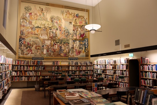 A well-lit library room with numerous bookshelves filled with books. In the center of the image, a large, colorful mural depicting various historical or cultural scenes covers the upper part of the wall. Several wooden tables and chairs are arranged neatly in the middle of the room. A modern light fixture hangs from the ceiling, casting warm light over the area.