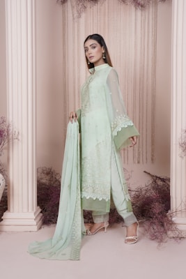 A woman is wearing a light pastel green traditional outfit with lace detailing and floral embroidery, standing gracefully between two white columns. Delicate dried flowers and fabric hang in the background, adding a whimsical touch to the scene. She holds the dupatta elegantly draped over her arm, and her expression is poised and serene.