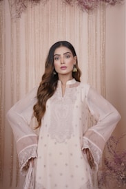 A woman stands confidently, wearing a traditional embroidered outfit in light shades. Her hair is styled in loose waves, and she accessorizes with gold earrings. The background features vertical hanging beads and dried floral accents, giving a subtle, elegant look.