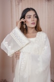 A woman with long brown hair is wearing a traditional, embroidered white outfit. The fabric features intricate patterns and delicate lace details. She holds one hand near her face, showcasing elegant earrings. The background consists of soft, cascading drapes in a neutral color.