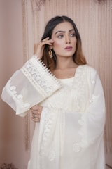 A woman with long brown hair is wearing a traditional, embroidered white outfit. The fabric features intricate patterns and delicate lace details. She holds one hand near her face, showcasing elegant earrings. The background consists of soft, cascading drapes in a neutral color.