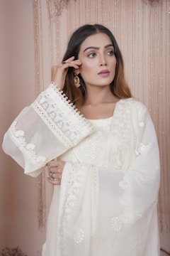 A woman with long brown hair is wearing a traditional, embroidered white outfit. The fabric features intricate patterns and delicate lace details. She holds one hand near her face, showcasing elegant earrings. The background consists of soft, cascading drapes in a neutral color.