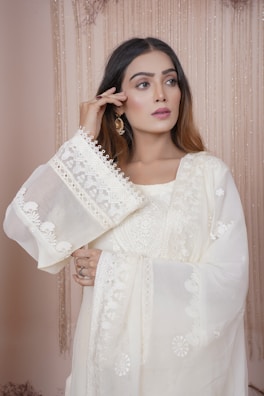 A woman with long brown hair is wearing a traditional, embroidered white outfit. The fabric features intricate patterns and delicate lace details. She holds one hand near her face, showcasing elegant earrings. The background consists of soft, cascading drapes in a neutral color.