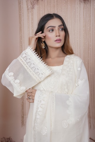 A woman with long brown hair is wearing a traditional, embroidered white outfit. The fabric features intricate patterns and delicate lace details. She holds one hand near her face, showcasing elegant earrings. The background consists of soft, cascading drapes in a neutral color.