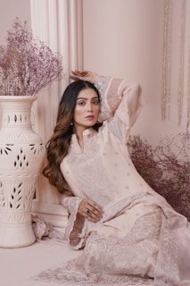 A woman is posed elegantly, wearing an intricately embroidered cream-colored traditional outfit with delicate lace sleeves. She is leaning against a decorative column with her hand gracefully positioned above her head. Beside her, there is an ornate white vase holding a bunch of dried, pinkish-purple flowers. The overall setting is soft and muted, with subtle textures and patterns enhancing the aesthetic.