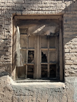 A weathered and broken window set in a rustic brick wall. The window has several missing and shattered glass panes, and the wooden frame is old and damaged. The bricks surrounding the window are rough and cracked, suggesting age and neglect.