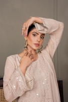 A woman is posing gracefully, wearing an elegant, embroidered beige outfit with intricate lace and mirror work. She accessorizes with large, ornate earrings and delicate jewelry. Her hand is placed artistically on her head, accentuating the design of her outfit.