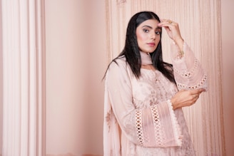 A softly lit photo of a woman wearing a flowing blush-pink anarkali dress with delicate gold embroidery, standing beside a vintage wooden doorway.