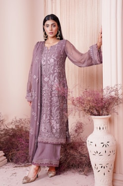 A woman dressed in an elegant, embroidered outfit in muted lilac tones stands gracefully against a backdrop with soft, draped fabric. Her attire is adorned with intricate floral patterns, adding a touch of sophistication. She wears statement earrings and stylish shoes, with her hair neatly styled. Decorative dried flowers and an ornate vase complete the aesthetically pleasing setting.