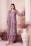 A woman dressed in an elegant, embroidered outfit in muted lilac tones stands gracefully against a backdrop with soft, draped fabric. Her attire is adorned with intricate floral patterns, adding a touch of sophistication. She wears statement earrings and stylish shoes, with her hair neatly styled. Decorative dried flowers and an ornate vase complete the aesthetically pleasing setting.