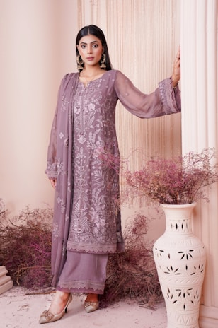 A woman dressed in an elegant, embroidered outfit in muted lilac tones stands gracefully against a backdrop with soft, draped fabric. Her attire is adorned with intricate floral patterns, adding a touch of sophistication. She wears statement earrings and stylish shoes, with her hair neatly styled. Decorative dried flowers and an ornate vase complete the aesthetically pleasing setting.