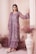 A woman dressed in an elegant, embroidered outfit in muted lilac tones stands gracefully against a backdrop with soft, draped fabric. Her attire is adorned with intricate floral patterns, adding a touch of sophistication. She wears statement earrings and stylish shoes, with her hair neatly styled. Decorative dried flowers and an ornate vase complete the aesthetically pleasing setting.