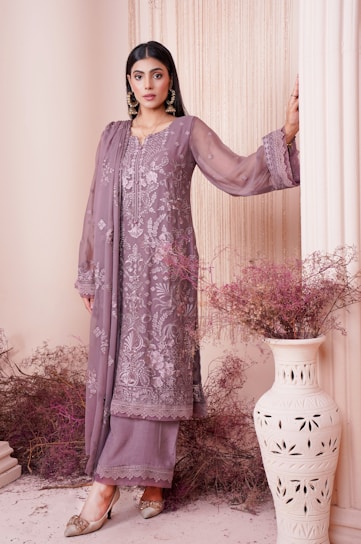 A woman dressed in an elegant, embroidered outfit in muted lilac tones stands gracefully against a backdrop with soft, draped fabric. Her attire is adorned with intricate floral patterns, adding a touch of sophistication. She wears statement earrings and stylish shoes, with her hair neatly styled. Decorative dried flowers and an ornate vase complete the aesthetically pleasing setting.