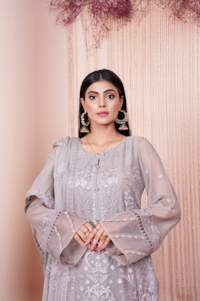 A woman wearing an intricately embroidered, floor-length, traditional outfit in a light lavender hue. Her dark hair is styled straight, and she accessorizes with large, golden hoop earrings. The background is soft and muted with cascading delicate fabrics and dried floral arrangements in shades of pink and purple.