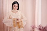 A woman is standing in an elegant pose, wearing a traditional embroidered outfit in soft beige tones with intricate detailing. She has long black hair and is adorned with statement earrings. Behind her is a pinkish-beige backdrop that includes a decorative column and some delicate floral arrangements.