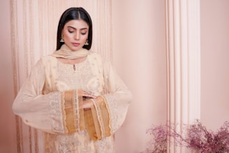 A woman is standing in an elegant pose, wearing a traditional embroidered outfit in soft beige tones with intricate detailing. She has long black hair and is adorned with statement earrings. Behind her is a pinkish-beige backdrop that includes a decorative column and some delicate floral arrangements.