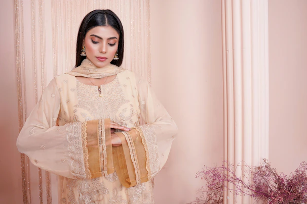 Elegant woman in a blush pink embroidered shalwar kameez standing against a soft beige backdrop.