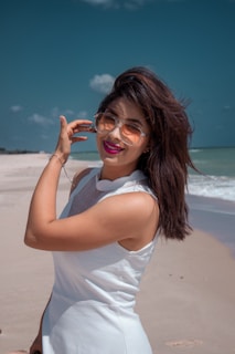 A woman with long dark hair wearing sunglasses and a sleeveless white dress stands on a sandy beach. She is smiling, with one hand touching her hair. The sky is clear with a few clouds, and the ocean is visible in the background.