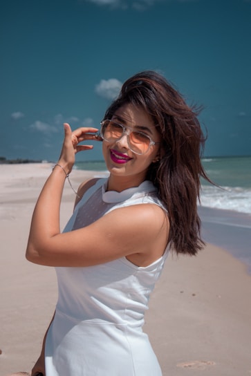 A woman with long dark hair wearing sunglasses and a sleeveless white dress stands on a sandy beach. She is smiling, with one hand touching her hair. The sky is clear with a few clouds, and the ocean is visible in the background.