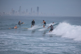 Several surfers are riding the waves in the ocean, with a distant city skyline visible on the horizon. The surfers appear to be skillfully maneuvering their boards through the water, and the scene is calm yet dynamic, with gentle waves rolling in.
