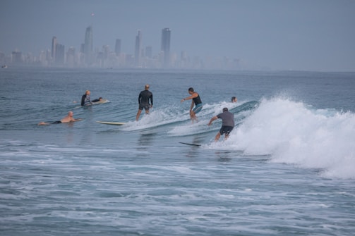 Several surfers are riding the waves in the ocean, with a distant city skyline visible on the horizon. The surfers appear to be skillfully maneuvering their boards through the water, and the scene is calm yet dynamic, with gentle waves rolling in.