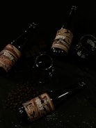 Three dark glass bottles with artistic labels are arranged on a surface surrounded by coffee beans and a few pieces of dark chocolate. There are also two empty glasses present beside the bottles.