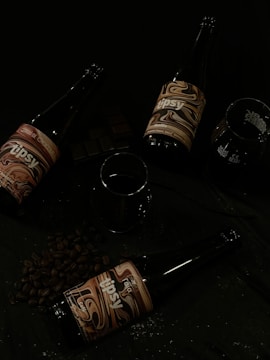 Three dark glass bottles with artistic labels are arranged on a surface surrounded by coffee beans and a few pieces of dark chocolate. There are also two empty glasses present beside the bottles.