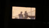 Blurred silhouettes of two people are visible, projected against a light background within a rectangular frame. The scene appears to be captured through a window or opening, creating a shadowy and mysterious effect.