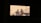 Blurred silhouettes of two people are visible, projected against a light background within a rectangular frame. The scene appears to be captured through a window or opening, creating a shadowy and mysterious effect.