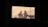 Blurred silhouettes of two people are visible, projected against a light background within a rectangular frame. The scene appears to be captured through a window or opening, creating a shadowy and mysterious effect.