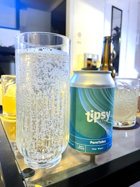 A clear glass filled with sparkling water is placed next to a blue and green can labeled 'tipsy PureTalus Hop Water' on a reflective table. In the background, there are additional glasses and a partially visible bottle, suggesting a setting of casual drinks.