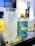 A clear glass filled with sparkling water is placed next to a blue and green can labeled 'tipsy PureTalus Hop Water' on a reflective table. In the background, there are additional glasses and a partially visible bottle, suggesting a setting of casual drinks.
