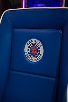 A blue padded chair featuring an embroidered emblem with a red lion and the words 'Rangers Football Club'. The background includes a pattern of dark blue and purple hues.