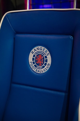 A blue padded chair featuring an embroidered emblem with a red lion and the words 'Rangers Football Club'. The background includes a pattern of dark blue and purple hues.