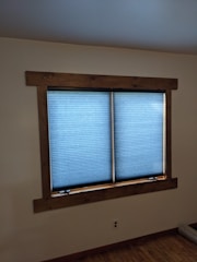 A window with blue blinds partially closed, set in a wooden frame, on an off-white wall. Below the window, there is an electrical outlet on the wall, and part of the wooden floor is visible.