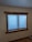 A window with blue blinds partially closed, set in a wooden frame, on an off-white wall. Below the window, there is an electrical outlet on the wall, and part of the wooden floor is visible.