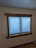 A window with blue blinds partially closed, set in a wooden frame, on an off-white wall. Below the window, there is an electrical outlet on the wall, and part of the wooden floor is visible.