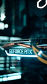 A close-up view of a computer graphics card with the text 'GEFORCE RTX' prominently displayed, featuring glowing lights and cables in a dimly lit environment.