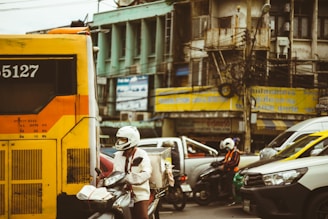 A busy urban street with a mix of vehicles including a bus, cars, and motorcycles. Traffic appears congested with a street background featuring old buildings and billboards. A person on a motorcycle is wearing a white helmet and jacket.