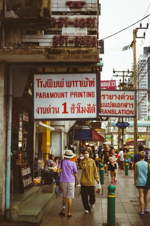 A busy urban street scene featuring people walking along a sidewalk lined with shops and signs in both English and Thai languages. The atmosphere appears lively with a mix of individuals casually strolling or engaged in conversation. Visible signs include those for a printing shop and translation services. Various utility poles and a cityscape can be seen in the background.